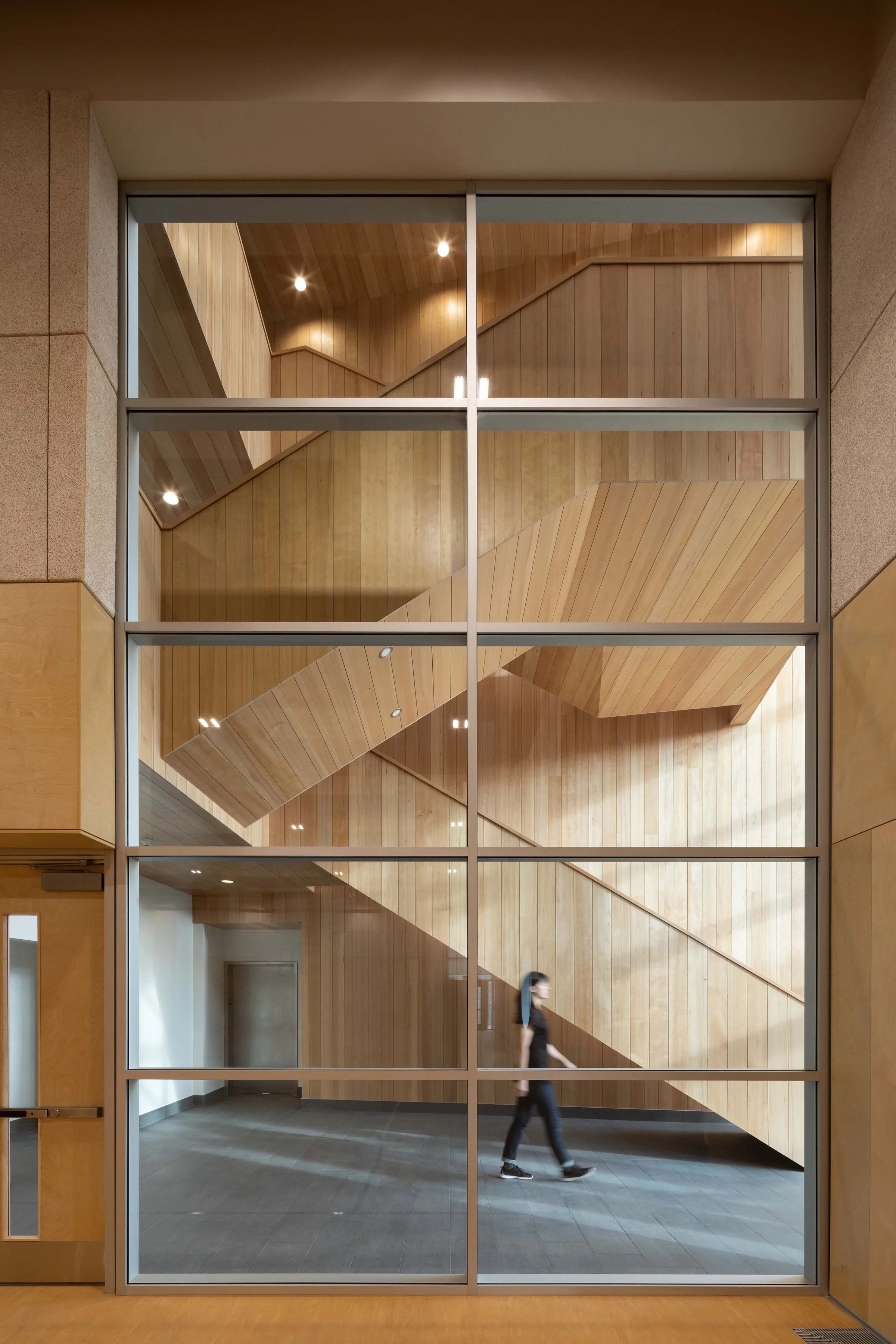 A full-height glazed wall revealing the locally sourced hemlock-lined stair, with a person crossing the lobby.